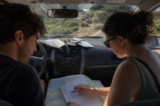 Two friends in a car stopped by Corsican roadside, reviewing paperwork and map with soft morning light through windows, surrounded by maquis vegetation.