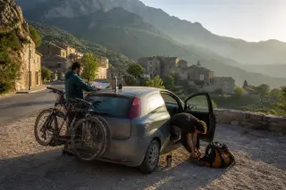 Two friends prepare for a day trip beside their parked car with bikes in a Corsican mountain village at sunrise, surrounded by hiking gear and old stone buildings.