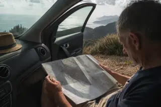Older traveler sitting in a parked car on a Corsican mountain road, reading a map with the sea and wild scenery visible through an open door in soft morning light.