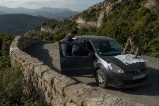 Two friends next to a small hatchback car on a mountain layby in Corsica, one reaching for a thermos, the other looking at a map on the hood, morning light, rugged road and landscape behind.