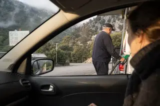 Mountain gas station in Corsica with a local attendant refueling a car, early morning light and steep hills in the background.