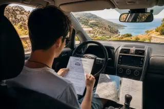 Driver in a rental car, paused on a Corsican mountain road, examining rental papers and a travel map, with a distant sea view through the windshield.