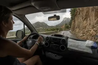 Woman driving on a narrow, winding Corsican mountain road after rain, recent rockfall visible, soft late afternoon light, natural candid atmosphere.