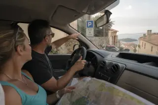 Corsican driver and tourist inside a car consult parking sign in narrow village street, morning light, candid travel moment