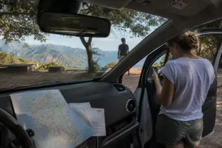 Interior of a rental car at a hillside Corsican rest stop with a woman checking her phone by the door and a man stretching, rugged coast and maquis visible under soft daylight.