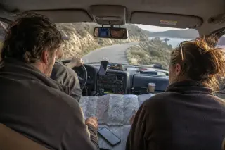View from the taxi back seat as two travelers consult a map, with a Corsican driver at the wheel on a winding mountain road, dashboard cluttered, morning light illuminating the scene.