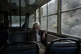 Elderly passenger quietly observing the mountain landscape from inside a Corsican bus, soft morning light streaming through a fogged window.