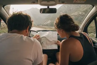 Two friends in a parked car on a Corsican mountain road, studying a map together in soft morning light, surrounded by rugged hills and maquis.