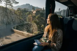 Young woman gazing out a bus window at Corsican mountain landscapes, early morning light creating subtle reflections inside the vehicle.