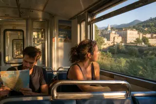 Passengers in a Corsican train carriage during golden hour, one reading a map, another looking out the window at a hillside village, with soft daylight and an authentic travel atmosphere.