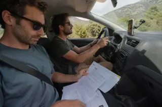 Two friends in a rental car, one driving and the other reviewing paperwork, on a winding Corsican road with maquis and hills outside, natural daylight, candid atmosphere.