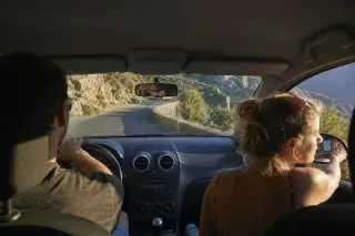 Two people in a car navigating a winding Corsican mountain road, focused on driving, seen from back seat with rugged scenery outside windows