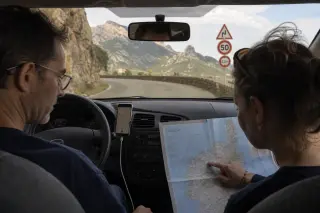 A man drives through Corsican mountains as a woman in the passenger seat traces a map, with road signs and rugged landscape outside, captured in soft morning light.