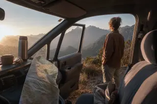 Unposed view from inside a car on a Corsican mountain pass, a woman stands outside gazing at the valley, soft morning light and travel items suggest a real journey pause.