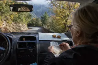 Older woman as passenger in car on a winding Corsican mountain road, unwrapping cheese, with map and sunglasses on dashboard, and natural autumn scenery outside.