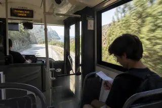 Corsican bus interior with driver focused on mountain road, young traveler checking ticket by window, coastal landscape visible outside.