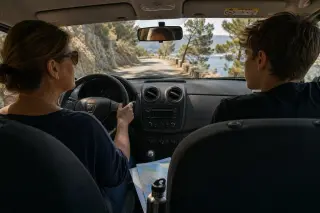Mother and teenage son in a car driving up a winding Corsican mountain road, softly lit by morning sun, with map and water bottle visible.