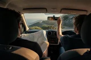 Two friends in a car pause on a Corsican mountain road, map open, natural morning light and scenic rugged landscape visible through windshield.