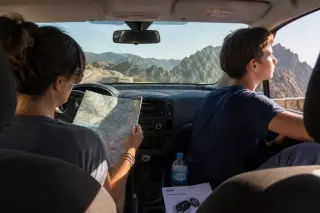 A woman behind the wheel of a parked car studying a map, with a teenage boy gazing through the passenger window at sunlit mountains, rental papers and car keys on the dashboard.