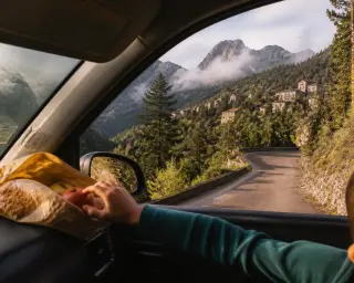 From inside a parked car, a child's hand reaches for fruit on the dashboard; view reveals a twisting Corsican mountain road, pine trees, stone houses, and soft morning light.