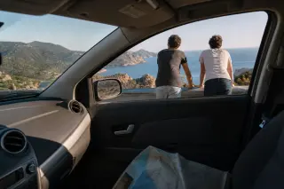 Two friends leaning at a scenic overlook near a coastal road in Corsica, viewed from inside a car with morning light and a rugged Mediterranean landscape.