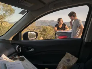 Two people at a roadside overlook in Corsica beside a compact rental car, interacting naturally with travel items and map, late afternoon light and maquis hills in background.