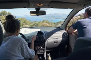 A couple in a parked car overlooking Corsican coastline, one reading a map, the other looking at the sea, in natural daylight.