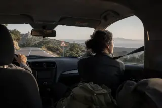 Two people in a car driving through Corsican mountains, seen from the back seat, with golden light, winding road, and sheep crossing sign ahead.