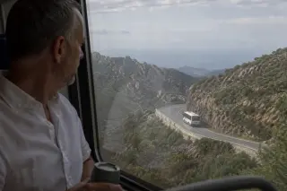 Passenger on a train looking out at a Corsican mountain road from a window, with a bus visible on the winding route, early morning light.