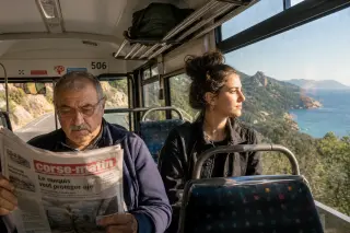 A Corsican bus interior with two people, sunlight streaming through the window, mountains and sea visible outside, calm and authentic atmosphere.