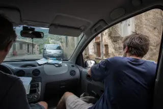 Two friends in a parked car in a Corsican mountain village, one checking paperwork on the dashboard, the other gazing out the window, photographed through the passenger seat with natural morning light and authentic ambiance.