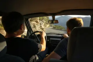 Corsican driver and friend navigating a sharp mountain turn, seen from the backseat, with golden afternoon light and a view of rough landscape through the car windows.