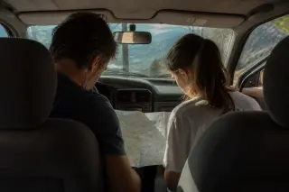 Father and teenage daughter inside a car, looking at a paper map together during a stop on a mountain road in Corsica, with natural golden light and scenic views outside the window.
