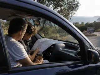 Couple in a rental car parked by a Corsican coastal road in morning light, consulting a map with a campsite sign and sea in the background.