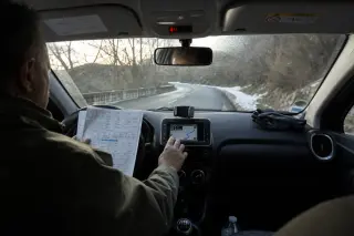 Conducteur dans une voiture arrêtée sur une route de montagne corse, consultant un GPS et une feuille météo, lumière hivernale douce, traces de neige et ambiance réaliste de voyage d’hiver