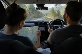 Young woman checks a paper map in a car as the driver navigates a winding tree-lined road in Corsica, morning light creating natural highlights, shot candidly from the back seat.
