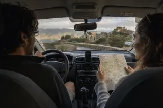 Couple in a rental car navigating a winding mountain road, with a map in hand and golden light illuminating a rugged Corsican landscape outside.