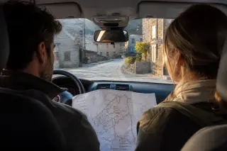 Two travelers in a parked car, looking at a map while quietly observing a peaceful Corsican village street at golden hour.