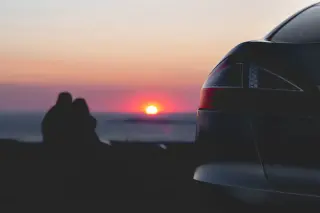 Voiture vue de l'arrière garée en face de la mer avec un couple en arrière plan qui regarde le coucher de soleil