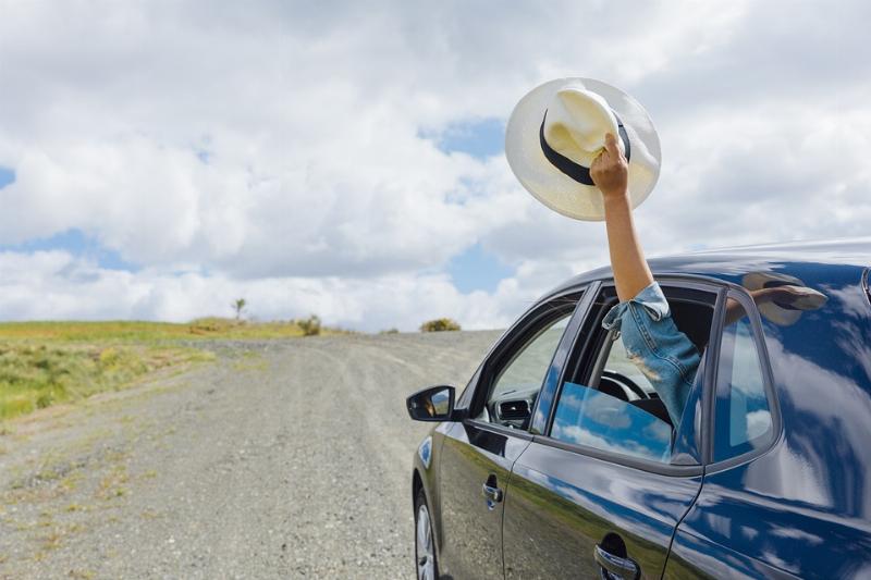 Un homme tend un chapeau à l'extérieur d'une voiture bleu qui roule sur un route déserte