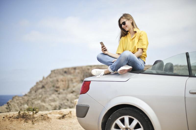 Jeune femme avec une chemise jaune regardant son téléphone et assise sur le coffre d'une voiture